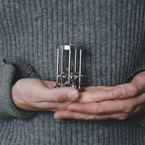 May include: A silver metal puzzle held in open hands. The puzzle is a cage-like structure with internal star-shaped pieces. The background is a grey knit sweater.