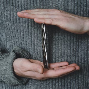 May include: A spiralled metal bar with a silver and dark brown finish is held between two hands. The hands are cupped around the bar, with the fingers and palms visible. The background is a grey jumper.