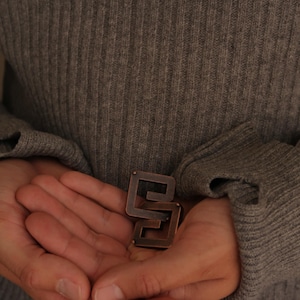 May include: A metallic, copper-toned puzzle piece featuring a geometric, interlocking square design. The object has a smooth, dark finish with visible edges and a distinct, angular shape, held in a cupped position against a grey ribbed fabric.