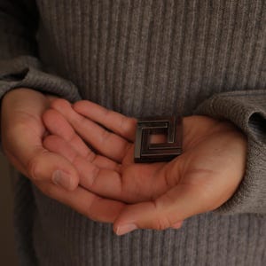May include: A small, square-shaped metal puzzle piece with a dark, gunmetal grey finish. The object features a geometric, interlocking design with a hollow centre, constructed from four distinct segments that form a continuous, closed loop.