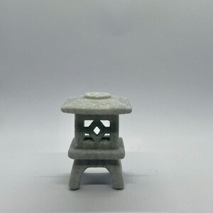 Könnte beinhalten: Eine kleine, graue japanische Gartenlaterne aus Stein. Die Laterne hat eine quadratische Ober- und Unterseite mit einem dekorativen rautenförmigen Ausschnitt im Mittelteil. Die Laterne ist eine Miniaturversion eines traditionellen Gartenelements.