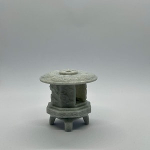 Puede incluir: Una pequeña linterna de piedra gris claro con una parte superior redonda y una base hexagonal. La linterna tiene una superficie texturizada y está sostenida por tres patas pequeñas. La parte superior tiene un agujero en el centro. Es un objeto decorativo.