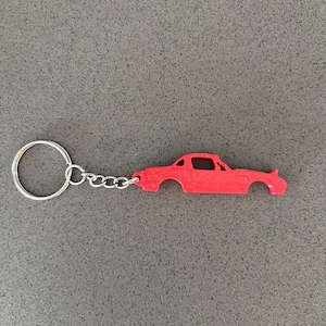 Pode incluir: Um porta-chaves vermelho brilhante com um recorte plano em forma de silhueta de carro desportivo. A forma do carro está presa a um anel circular de metal prateado padrão por uma corrente curta de metal prateado. Mostra o perfil lateral.