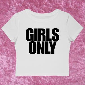 May include: White crop top with the text "GIRLS ONLY" in bold black letters. The shirt has short sleeves and a crew neck. The background is a pink, textured surface.
