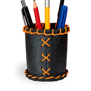 May include: A black pen holder with orange stitching and decorative X's, holding pens and pencils. The holder has a textured surface and a decorative orange trim around the top and bottom. The pencils are red and yellow.