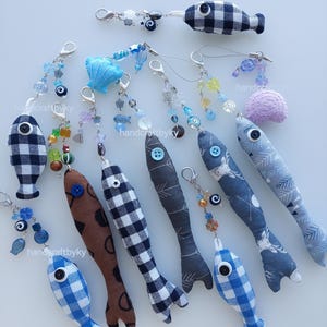 Peut inclure: Une collection de porte-clés faits à la main en forme de poisson, avec divers motifs et couleurs, dont du vichy bleu et blanc, du marron à pois et du gris à flèches. Chaque porte-clés est orné d'un bouton en guise d'œil et de perles décoratives.