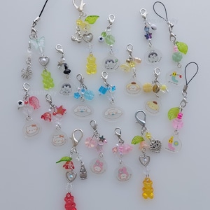 May include: A collection of various phone charms featuring metal lobster clasps, colourful acrylic beads, small metal charms, and translucent circular pendants with printed cartoon characters, including bears, animals, and floral motifs.