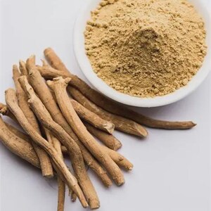 Op de afbeelding: Een close-up van ashwagandha-wortel en -poeder. De afbeelding toont een stapel lichtbruine ashwagandha-wortels naast een witte kom gevuld met een fijn, bruin poeder. De achtergrond is een effen wit oppervlak.