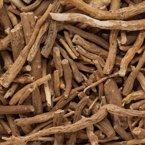 Op de afbeelding: Close-up van stukjes ashwagandha-wortel. De stukjes zijn verschillend van grootte en vorm, met een lichtbruine kleur en een ruwe textuur. Sommige stukjes hebben een lichtere, bijna witte buitenlaag. De afbeelding is goed belicht.
