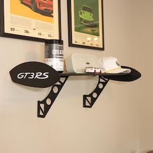 May include: A black wall-mounted shelf featuring side panels shaped like car spoilers with the text GT3RS in white. The shelf supports a black container, a light gray rectangular speaker, a small white toy car, and a white baseball cap.