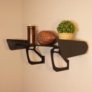 May include: A black metal wall shelf with a unique design, holding a brown leather football, a small potted plant, and a wooden container. The shelf is mounted on a beige wall, creating a modern display.
