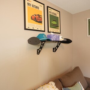 May include: Two framed posters with the text "911 GT3RS" and images of a red and green sports car. A black shelf holds a teal cap, a white cap, and a box of tissues. The shelf is mounted on a beige wall.