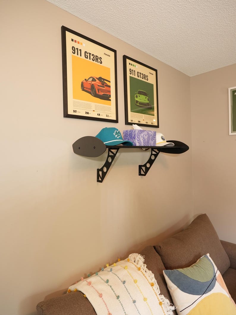 May include: Two framed posters with the text "911 GT3RS" and images of a red and green sports car. A black shelf holds a teal cap, a white cap, and a box of tissues. The shelf is mounted on a beige wall.