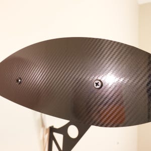 May include: A black carbon fiber spoiler with a textured weave pattern. Two screws secure the spoiler to a black metal support structure. The spoiler has a curved shape and is designed for automotive use.