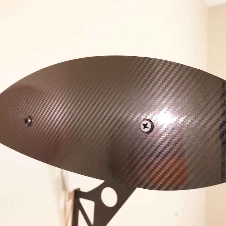 May include: A black carbon fiber spoiler with a textured weave pattern. Two screws secure the spoiler to a black metal support structure. The spoiler has a curved shape and is designed for automotive use.
