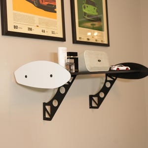 May include: A black metal wall shelf with a white oval accent piece. The shelf holds a small white toy car, a speaker, and other decorative items. Two framed posters hang above the shelf.
