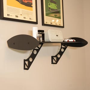 May include: Black metal wall shelf with a unique wing-like design. The shelf holds a white speaker, a small toy car, and other decorative items. The shelf is mounted on a light-colored wall, with framed posters above.