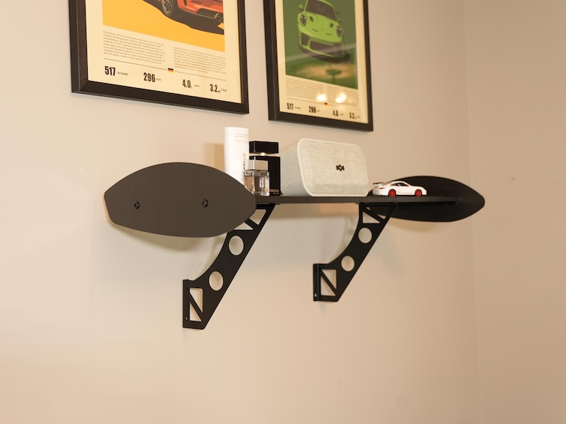 May include: Black metal wall shelf with a unique wing-like design. The shelf holds a white speaker, a small toy car, and other decorative items. The shelf is mounted on a light-colored wall, with framed posters above.