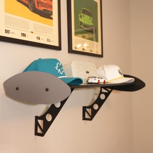 May include: A black wall shelf with a carbon fiber-style front panel, holding a teal baseball cap with a white logo, a beige box, a white cap, and a small toy car. Two framed posters are visible in the background.