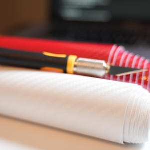 May include: A roll of white textured vinyl material with a black and yellow utility knife and a red roll of material in the background. The vinyl has a subtle pattern. The knife is in focus, with the blade exposed.