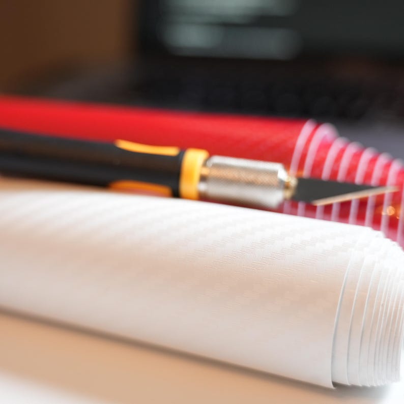 May include: A roll of white textured vinyl material with a black and yellow utility knife and a red roll of material in the background. The vinyl has a subtle pattern. The knife is in focus, with the blade exposed.