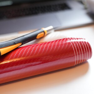 May include: A roll of glossy red carbon fiber vinyl with a black and orange utility knife. The vinyl has a woven pattern and is partially unrolled. A laptop is visible in the background.