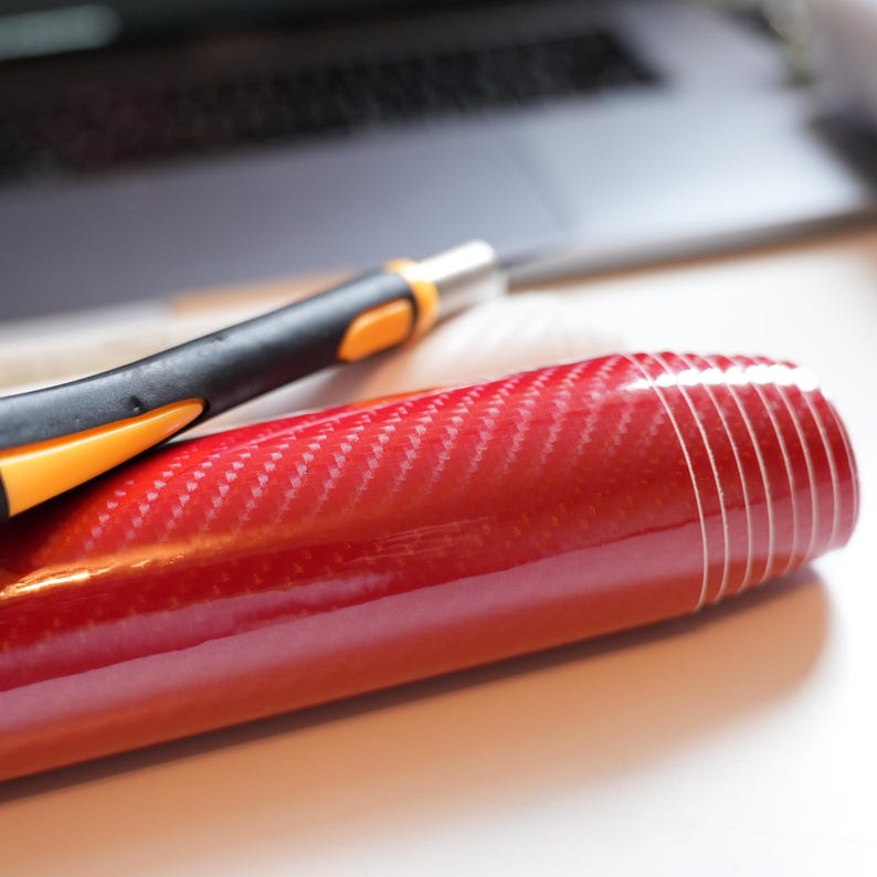 May include: A roll of glossy red carbon fiber vinyl with a black and orange utility knife. The vinyl has a woven pattern and is partially unrolled. A laptop is visible in the background.