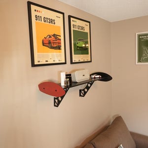 May include: Two framed posters with the text "911 GT3RS" and images of sports cars hang on a beige wall. A black shelf with a red accent holds a speaker, small car model, and other items. A brown sofa with decorative pillows is in the foreground.