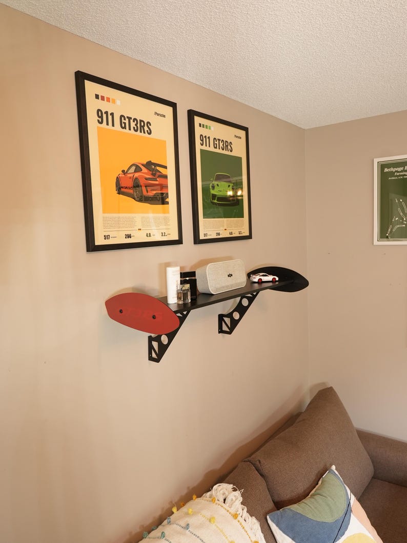 May include: Two framed posters with the text "911 GT3RS" and images of sports cars hang on a beige wall. A black shelf with a red accent holds a speaker, small car model, and other items. A brown sofa with decorative pillows is in the foreground.