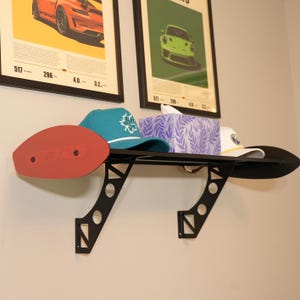 May include: A black metal shelf in the shape of a car spoiler, mounted on a wall. The shelf holds a red oval piece, a teal cap with a white maple leaf, a tissue box, and a white cap. Two framed car posters are in the background.