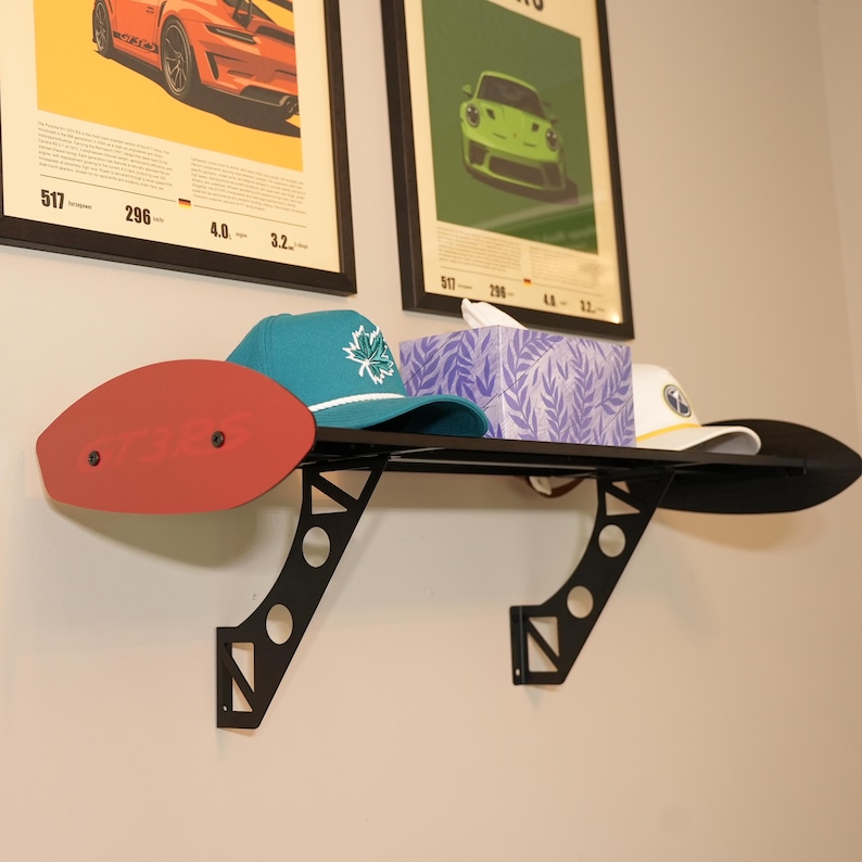 May include: A black metal shelf in the shape of a car spoiler, mounted on a wall. The shelf holds a red oval piece, a teal cap with a white maple leaf, a tissue box, and a white cap. Two framed car posters are in the background.