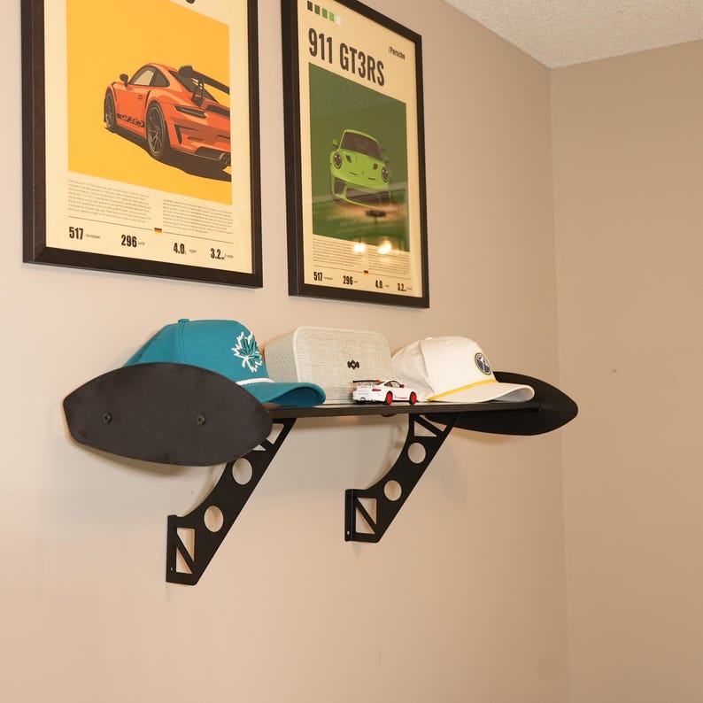 May include: A black metal shelf with a car spoiler design, mounted on a beige wall. The shelf holds a blue baseball cap with a white logo, a white cap with a yellow brim, a small white box, and a toy car.