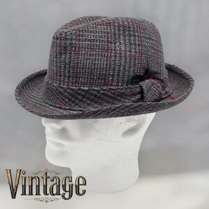 May include: A grey fedora hat with a plaid pattern in shades of grey and burgundy. The hat has a small bow detail on the side and a classic brim. The word "Vintage" is displayed in gold script at the bottom.