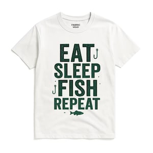 Puede incluir: Camiseta blanca con el texto "EAT SLEEP FISH REPEAT" en verde oscuro, junto con un pequeño gráfico de pez. La camiseta tiene cuello redondo y mangas cortas. La marca "FABRIC WEAR" está impresa en el cuello.