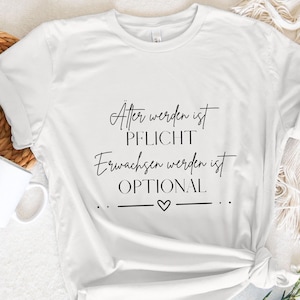 Puede incluir: Camiseta blanca con el texto "Älter werden ist PFLICHT, Erwachsen werden ist OPTIONAL" en escritura negra. La camiseta tiene un dobladillo anudado y está diseñada sobre una superficie blanca con una taza y vegetación.