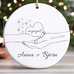 Könnte beinhalten: Runder Weihnachtsbaumschmuck aus weißer Keramik mit goldenem Aufhänger. Das Design zeigt eine schwarze Strichzeichnung einer Hand, die ein Herz mit dem Wort "engaged" hält. Das Datum "25.02.2026" und die Namen "Anna + Björn" sind ebenfalls aufgedruckt.