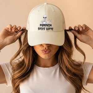 Pode incluir: Um boné trucker cor de creme com parte traseira em rede e painel frontal sólido. A frente apresenta um gráfico preto de um esqueleto segurando uma chávena de café e fazendo um gesto com a mão, com o texto PUMPKIN SPICE LATTE.