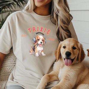 May include: A beige short-sleeve t-shirt featuring a graphic of a beagle puppy wearing headphones. The text TRIXIE is printed in an arched font above the puppy, with EST on the left and 2026 on the right in a matching orange and pink color scheme.