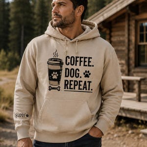 Könnte beinhalten: Ein beigefarbener Kapuzenpullover mit einer schwarzen Grafik, die einen Kaffeebecher mit Pfotenabdruck, den Text "COFFEE. DOG. REPEAT." und ein Knochen-Symbol zeigt. Auf dem rechten Ärmel steht der Name "BUDDY" neben einem kleinen Pfotenabdruck.