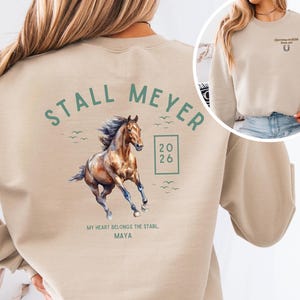 Puede incluir: Sudadera beige con ilustración de caballo. El diseño incluye un caballo marrón, las palabras "STALL MEYER", los números "20 26" en un cuadrado y el texto "MY HEART BELONGS THE STABL, MAYA".