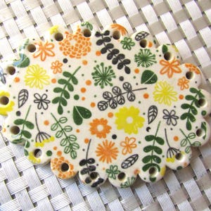 May include: A ceramic oval-shaped bead with a scalloped edge. The bead is white with a colorful floral pattern in yellow, orange, green, and black. Small holes are evenly spaced around the edge.