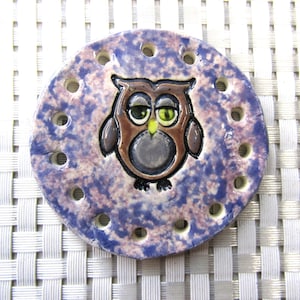 May include: A ceramic owl stitch marker with a purple and pink speckled glaze. The owl has brown feathers, a gray belly, and yellow eyes. The marker has holes around the edge for holding yarn.