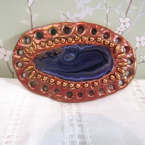 May include: An oval brooch featuring a central dark blue agate stone with visible banding. The stone is surrounded by a textured, metallic copper-coloured frame decorated with small gold-toned beads and a series of evenly spaced circular cutouts.