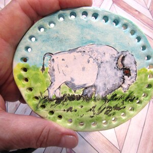 May include: An oval ceramic plaque featuring a hand-painted white bison standing on a green grassy field against a light blue sky. The plaque has a series of evenly spaced holes punched around the entire perimeter for threading or attachment.