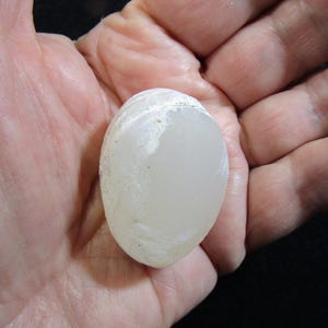 Puede incluir: Una piedra lisa, blanca y ovalada con una textura ligeramente rugosa. La piedra es opaca y tiene un color blanco lechoso.