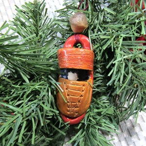 May include: A handmade ceramic Christmas ornament shaped like a boot with a red top, brown laces, and a white interior. The ornament is hanging from a red string and a brown bead.