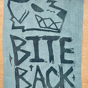 May include: A rectangular fabric patch in a muted sage green color featuring a black screen-printed graphic of a stylized dog head with bared teeth above the bold, distressed black text that reads BITE BACK, accented by small four-pointed stars.