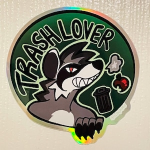 Pode incluir: Um autocolante holográfico redondo com um guaxinim de desenho animado e o texto "TRASH LOVER" num círculo verde. O guaxinim tem um padrão de pelo preto e cinzento, olhos vermelhos e um caixote do lixo.