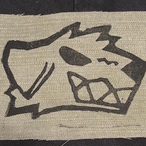 May include: A rectangular beige fabric patch featuring a bold black line drawing of a stylized dog head with a jagged ear and visible teeth. The patch is stitched onto a black fabric background with visible frayed edges along the perimeter.