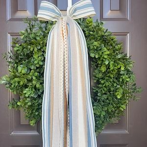 May include: A green boxwood wreath with a blue and white striped bow and cascading ribbons. The ribbons include lace, gingham, and solid colors of blue, beige, and white. The wreath hangs on a brown door.